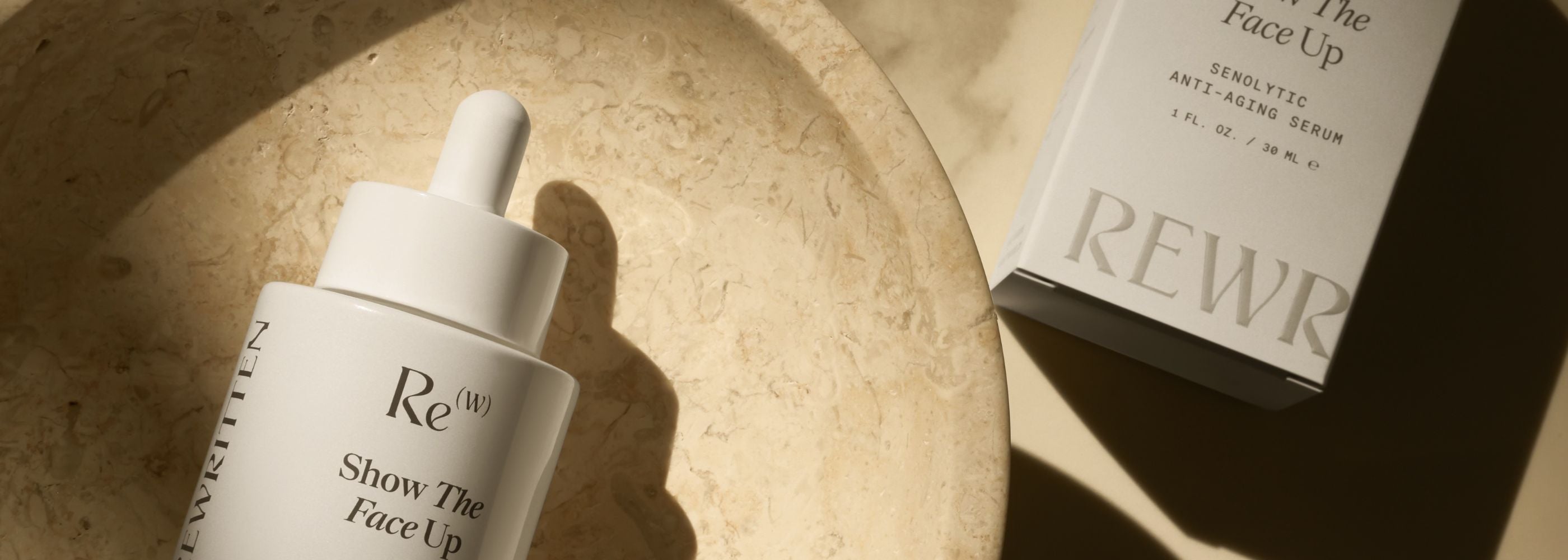 Flatlay of Rewritten Show The Face Up senolytic anti-aging serum bottle and packaging box placed on a stone surface in warm natural light